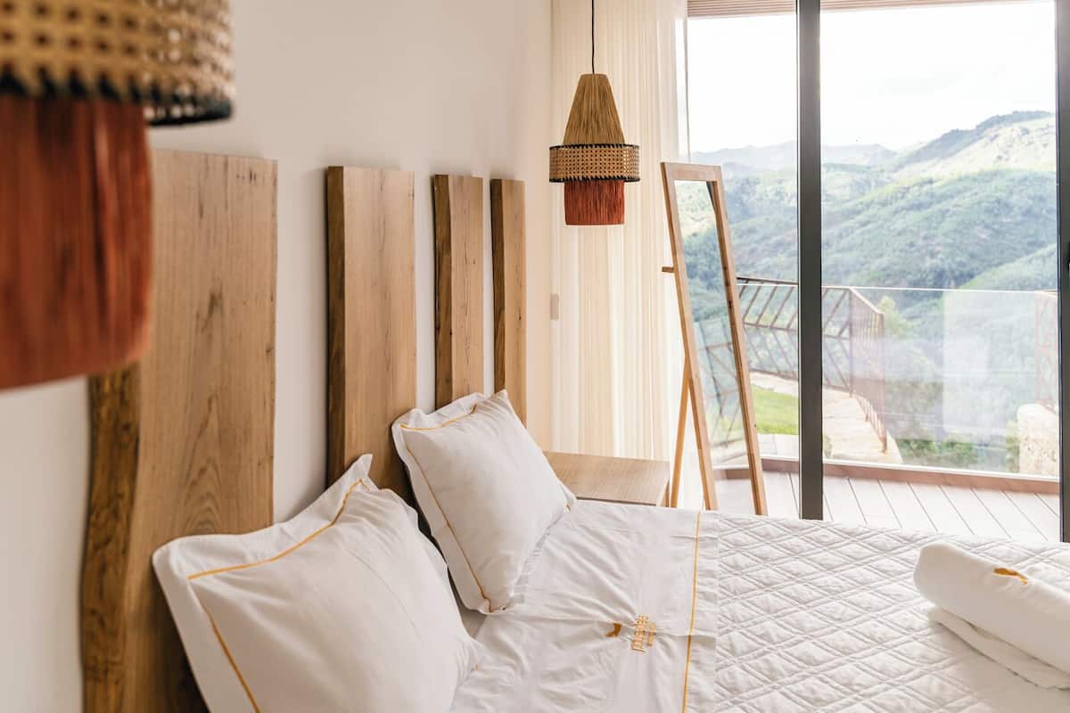 Une chambre à coucher avec une tête de lit en bois, un lit blanc avec des oreillers assortis, un luminaire suspendu et une vue sur un paysage montagneux par de grandes fenêtres.