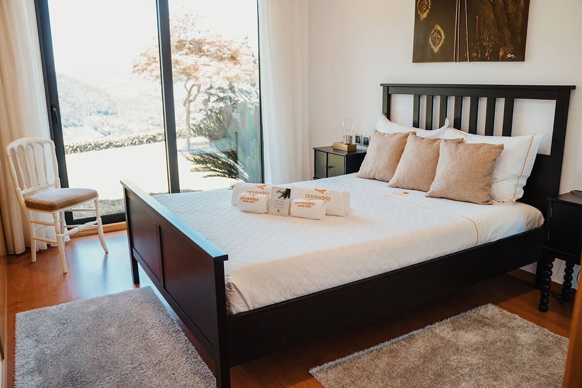 Un lit bien fait, avec des oreillers et une couverture, une chaise, une table de chevet avec une lampe, et une grande fenêtre donnant sur des arbres et des collines.