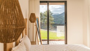 A bedroom with a large bed, a view of a balcony with a wooden deck, and a mountain landscape.