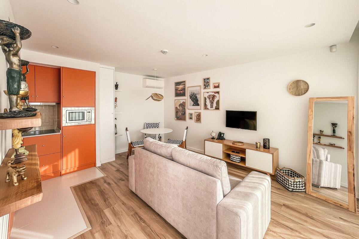 Un salon moderne avec un canapé, un coin repas comprenant une table et des chaises, une cuisine avec des meubles orange et un téléviseur fixé au mur.