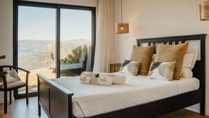 A bedroom with a large bed, a chair, a nightstand, and a view of the outdoors.