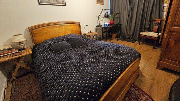 Une chambre à coucher bien rangée, avec un lit en bois, une petite table de chevet avec une lampe, une chaise et un bureau agrémenté d’une plante.