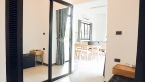 A modern living room with a black leather sofa, a sliding glass door leading to a dining area, and a kitchen visible through an open door. - Issara Private Pool Villa Khaoyai A (Pak Chong)