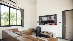 A modern living room with a large flat-screen TV mounted on the wall, a brown leather sofa, a small wooden side table, and a door to the right. - Issara Private Pool Villa Khaoyai A (Pak Chong)