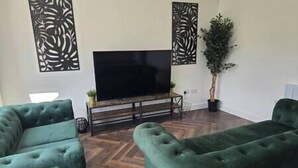 A living room with a flat-screen TV on a wooden stand, two green sofas, and decorative wall panels. - Welcome to our centrally-located 4-bedroom detached new build home in Stafford (Stafford)