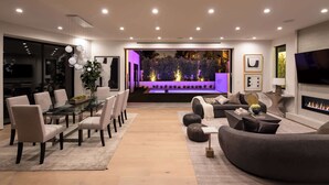 A modern living room with a dining area, a fireplace, and a view of a garden at night.