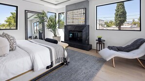 A modern bedroom with a large bed, a fireplace, a seating area with a chair, and large windows offering a view of the outdoors.