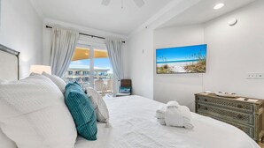 Et hotelværelse med en seng, en kommode, et maleri af en strand og udsigt til en bygning gennem vinduet.