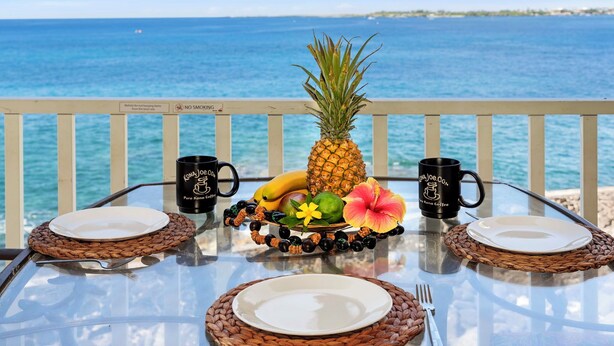 A table set with plates, cutlery, a pineapple, bananas, a flower, and two coffee mugs with a view of the ocean.