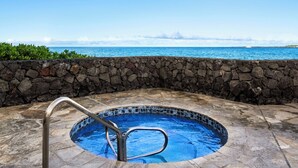 A stone-walled hot tub with a metal ladder, surrounded by stone tiles, overlooking a clear blue ocean.