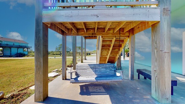 Eine Pergola aus Holz mit Betonboden, umgeben von einer Rasenfläche und einem Gebäude im Hintergrund.