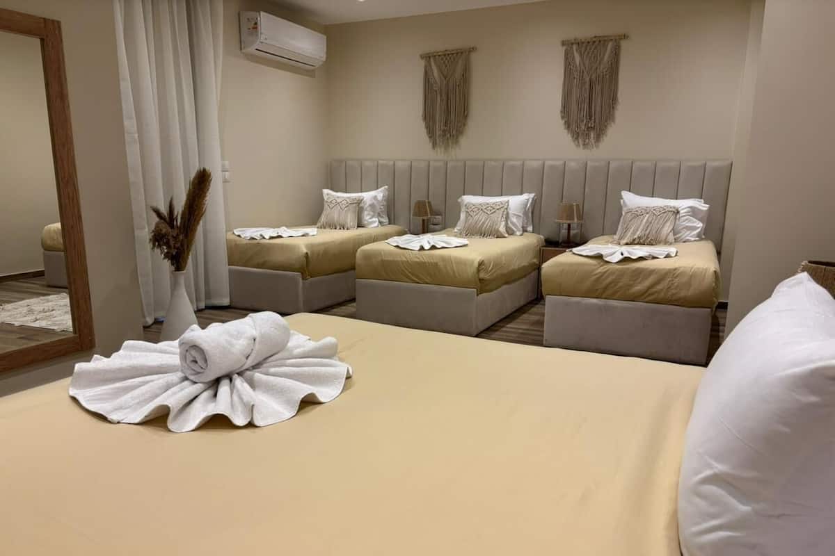 A hotel room with three beds, each with a beige bedspread and white pillows. There is a large mirror on the left wall, a wall-mounted air conditioner, and a decorative macrame hanging above the beds.