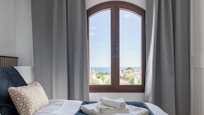 Ein Schlafzimmer mit Bett, grauen Vorhängen und einem Fenster mit Blick auf das Meer und Gebäude.