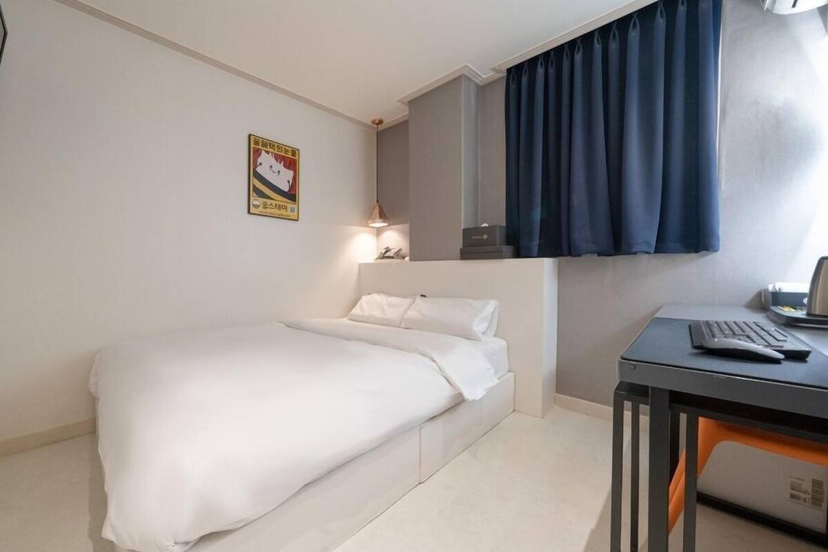 Quarto de hotel pequeno com uma cama de solteiro, uma escrivaninha com computador, uma cadeira, TV presa à parede e um cartaz na parede.