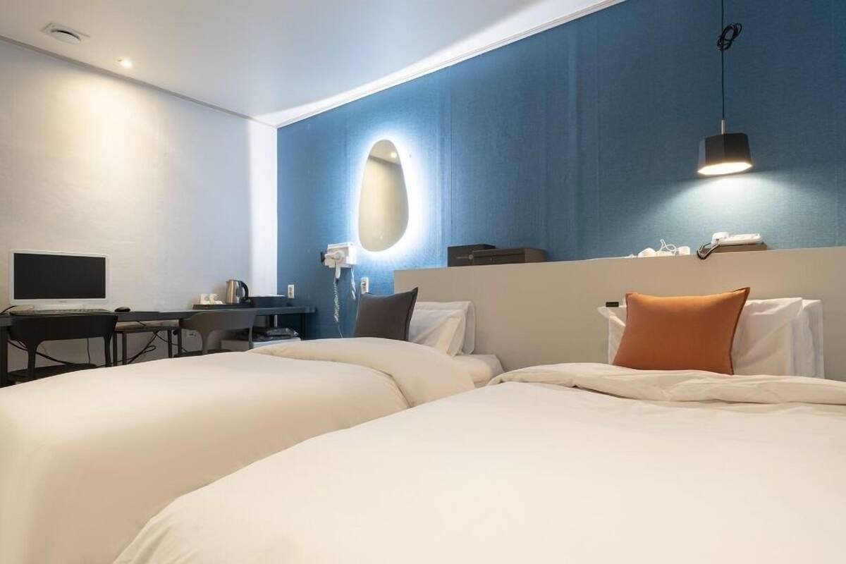 Quarto de hotel com duas camas, uma escrivaninha com computador, telefone e luminária fixa na parede.