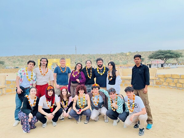A group of people posing in a sandy outdoor area with a stone wall and a clear sky in the background. - Osaka Desert Safari Camp (Jaisalmer)
