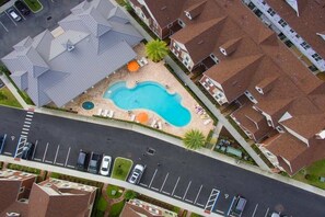 An aerial view of a residential area with a swimming pool surrounded by houses, a parking lot, and a road.