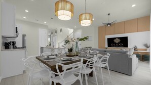Una cocina moderna con armarios blancos, un comedor con mesa de madera y sillas blancas, y una sala de estar con sofá gris y televisor montado.