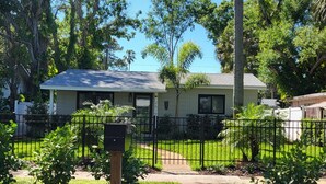 A residential house with a paved driveway, a mailbox, and a fenced yard with various trees and shrubs.