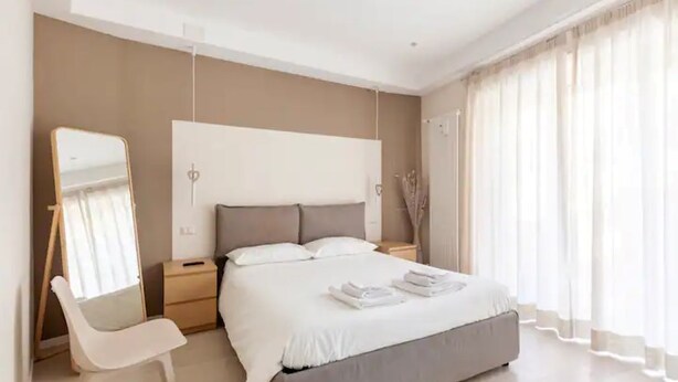 A modern bedroom with a large bed, a bedside table, a chair, a mirror, and sheer curtains. - Charming stay in Rome: Luxury apartment one step from St. Peter's Basilica (Roma)