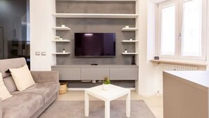 A modern living room with a flat-screen TV mounted on a grey wall, a grey sofa, a white coffee table, and a window with white shutters. - Charming stay in Rome: Luxury apartment one step from St. Peter's Basilica (Roma)