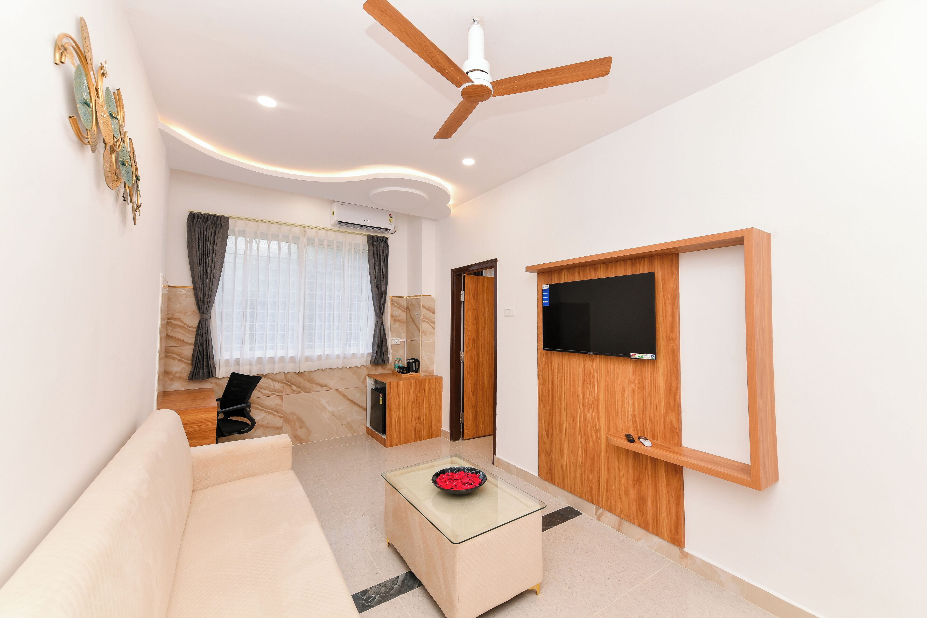 A modern living room with a sofa, a glass coffee table, a ceiling fan, a flat-screen TV mounted on the wall, and a door leading to another room.