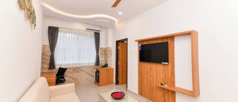 A modern living room with a sofa, a glass coffee table, a ceiling fan, a flat-screen TV mounted on the wall, and a door leading to another room.