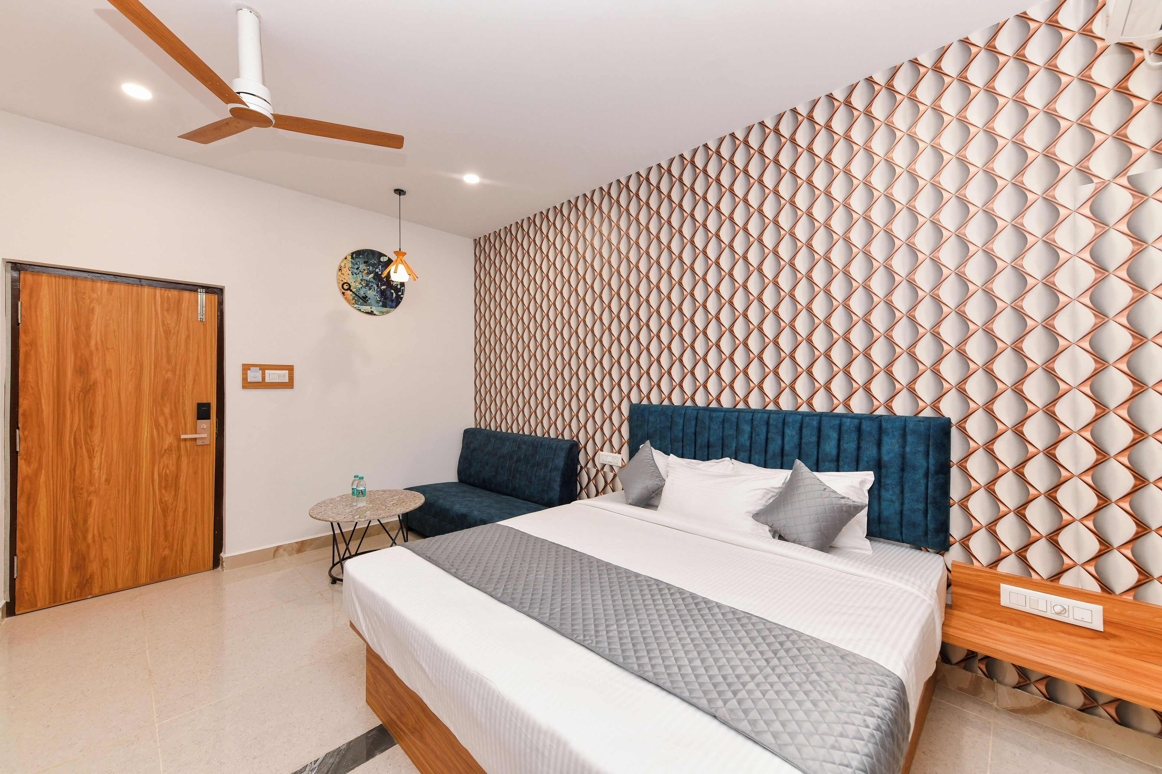 A modern bedroom with a wooden door, a round table, a bed with a blue headboard, and a patterned wall.