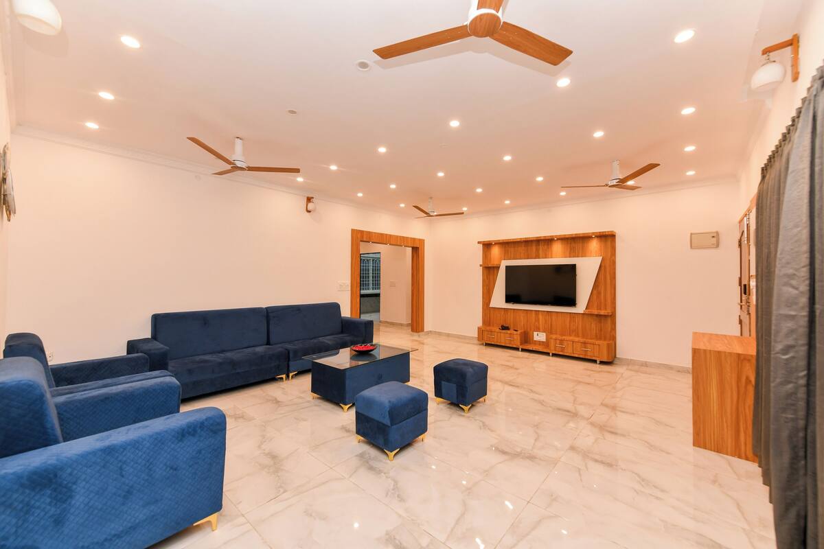 A spacious living room with a sectional sofa, ottomans, a coffee table, a flat-screen TV mounted on a wooden wall, ceiling fans, and recessed lighting.