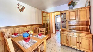 Un comedor con una mesa de madera preparada para cuatro personas, rodeada de sillas. Se ve una cocina al fondo con armarios de madera y un lavabo.
