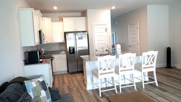 Un espacio habitable compacto con kitchenette, un área de comedor con sillas blancas y un sofá gris con cojines estampados.
