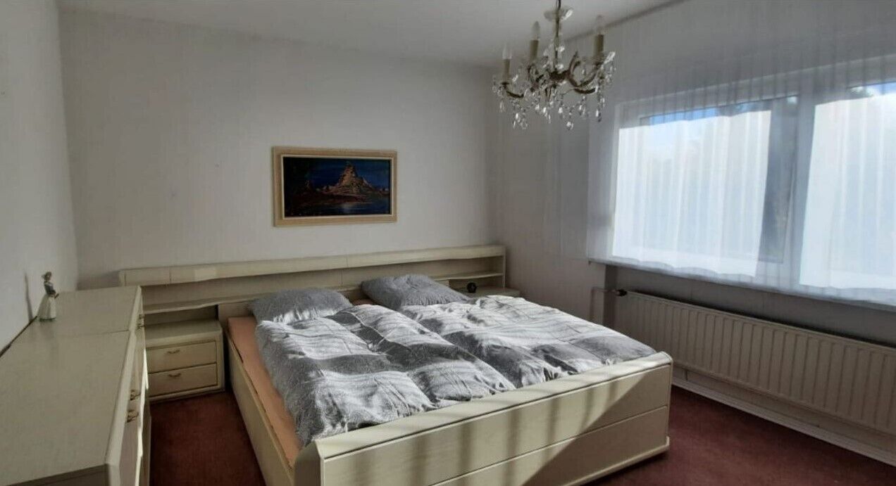 A bedroom with a bed, a nightstand, a dresser, a chandelier, a window with curtains, and a painting on the wall.
