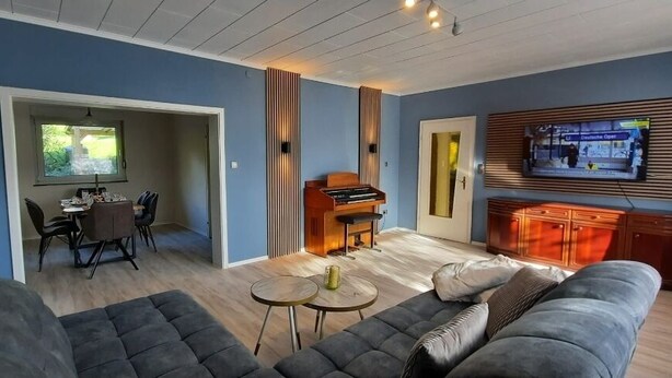 Un saló modern amb un piano, una televisió penjada en un panell de fusta, i una zona de menjador amb taula i cadires.