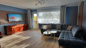 Un saló modern amb un sofà modular, un televisor muntat sobre un panell de fusta, una taula rodona petita i una finestra gran amb cortines.