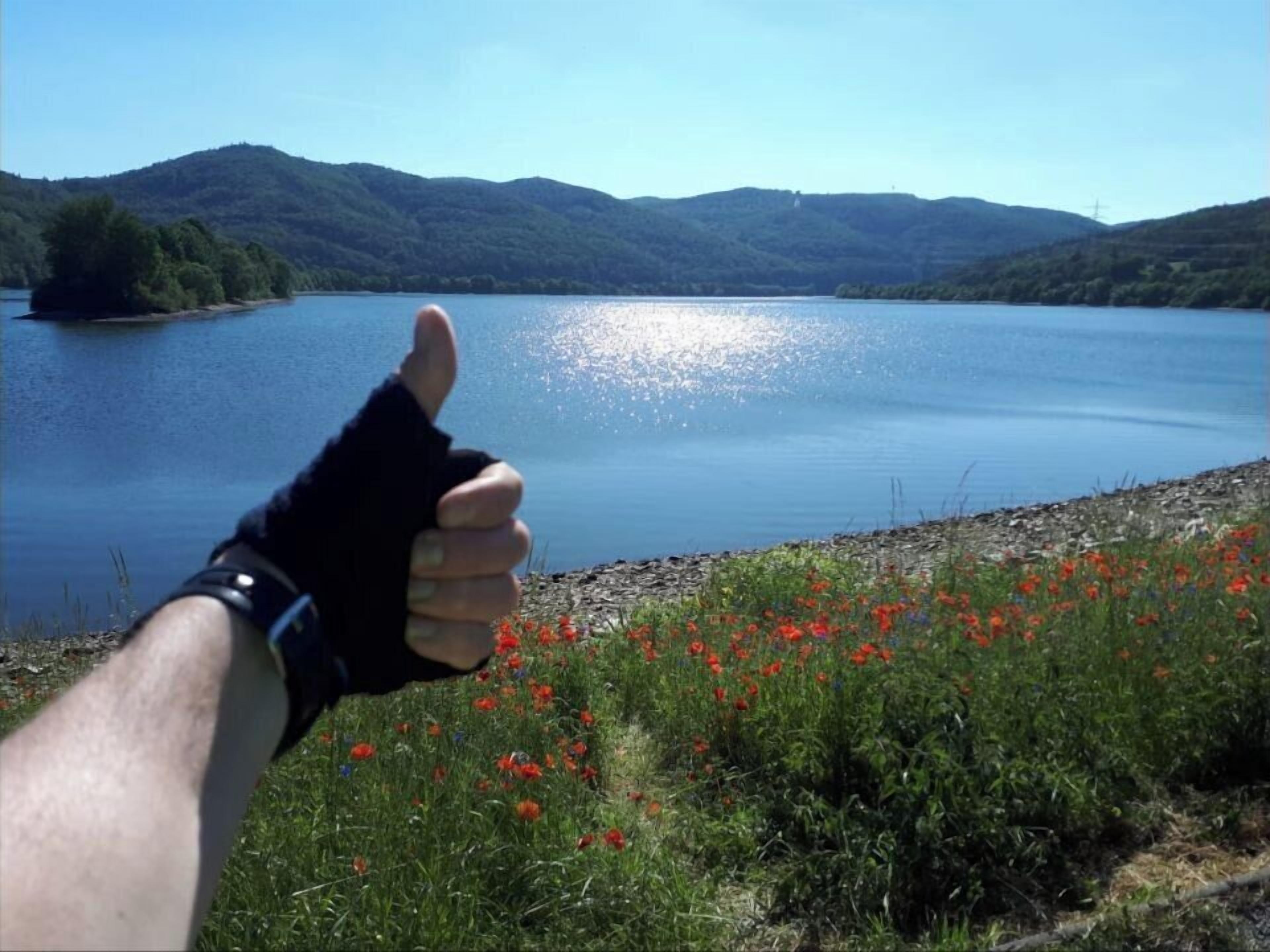 Eine Hand zeigt Daumen hoch, im Hintergrund ein See und Berge.