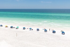 A beach with white sand and clear turquoise water. There are several umbrellas and people relaxing on the beach.