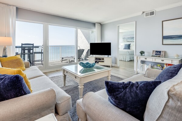 A spacious living room with a large window, a flat-screen TV, and a balcony with outdoor furniture.