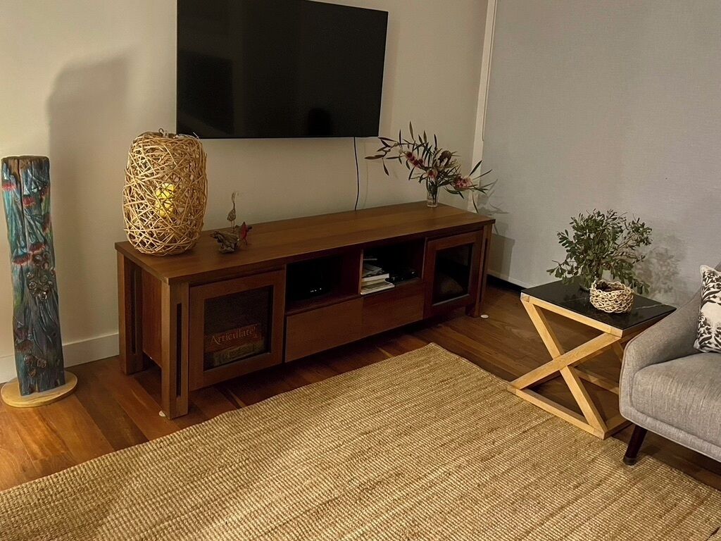Een woonkamer met een houten tv-meubel, een geweven lamp, een kleine tafel met een plant en een grijze bank.