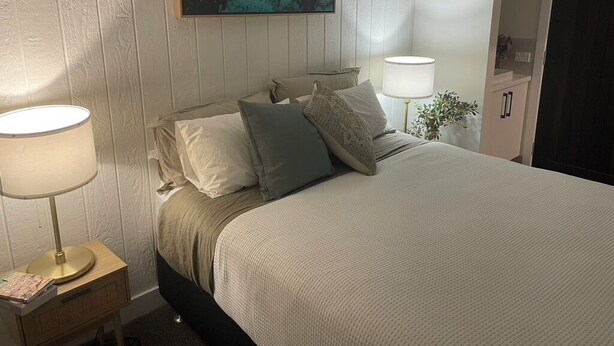 Una camera da letto con un letto, due abat-jour, un comodino, un quadro appeso al muro e una porta.