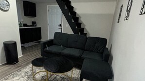 Un salon avec un canapé d’angle gris foncé, un pouf assorti et une table basse avec un cadre doré. On y trouve un escalier menant à un autre niveau, un coin cuisine avec des meubles sombres et une porte blanche.