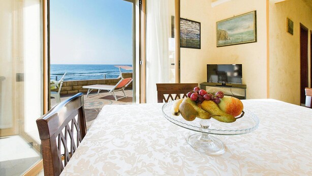 Un comedor con una mesa preparada para dos personas, un frutero sobre la mesa, un televisor en un mueble bajo y vistas al mar a través de una puerta abierta.