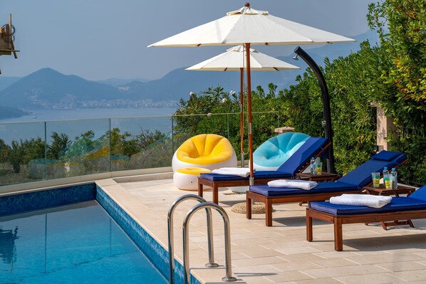Un espace aménagé au bord de la piscine, avec des chaises longues, des parasols et une vue dégagée sur la mer et les montagnes.