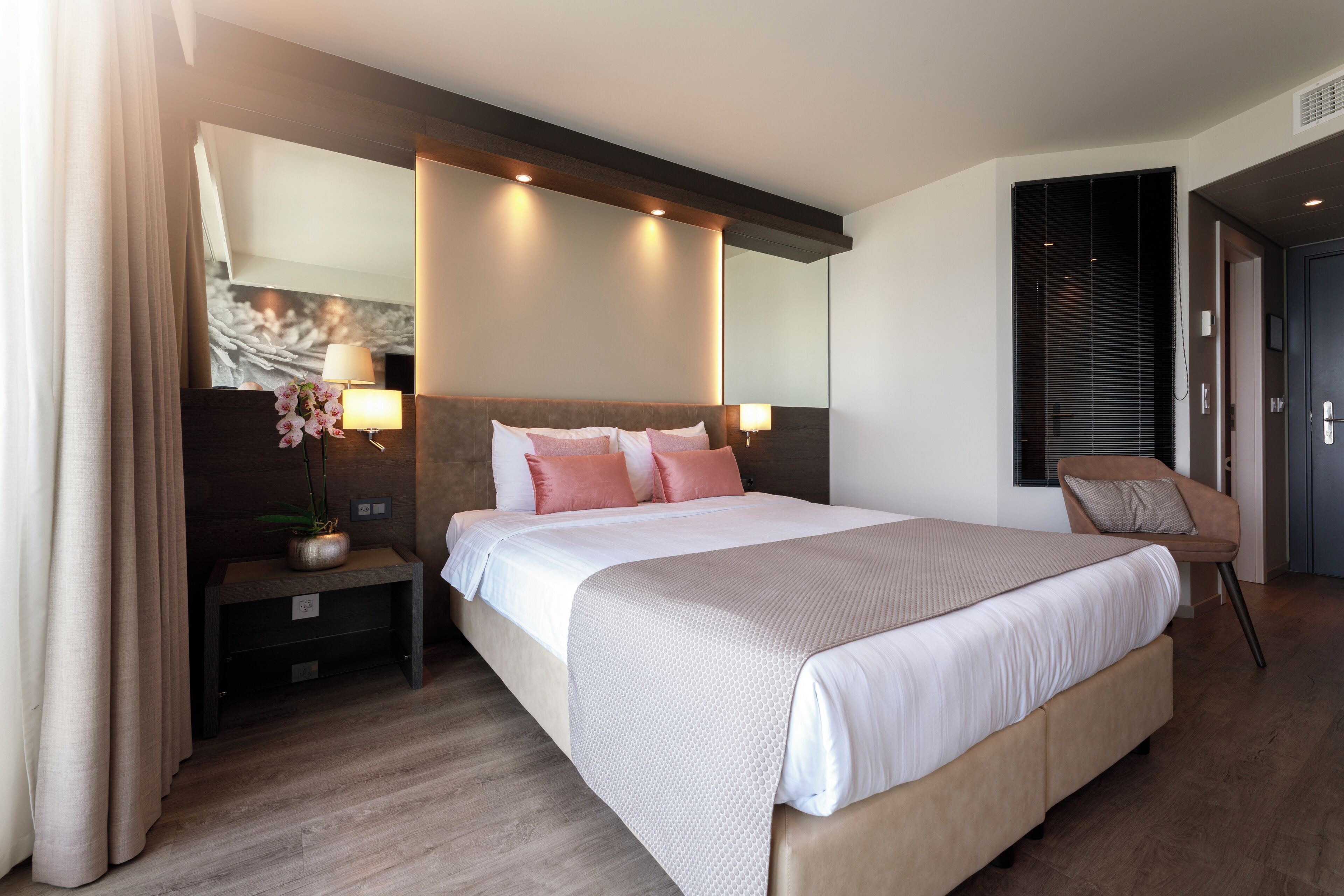 A modern hotel room with a large bed, bedside lamps, a nightstand, a chair, and a large mirror.