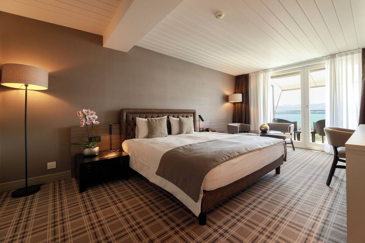 A hotel room with a large bed, a nightstand, a lamp, a chair, and a view of the sea.