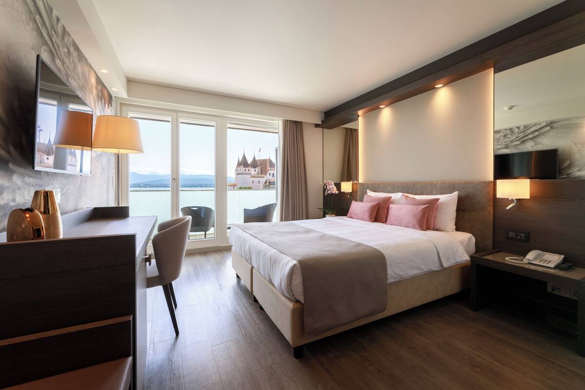 A modern hotel room with a large bed, a desk, and a view of a castle through the window.