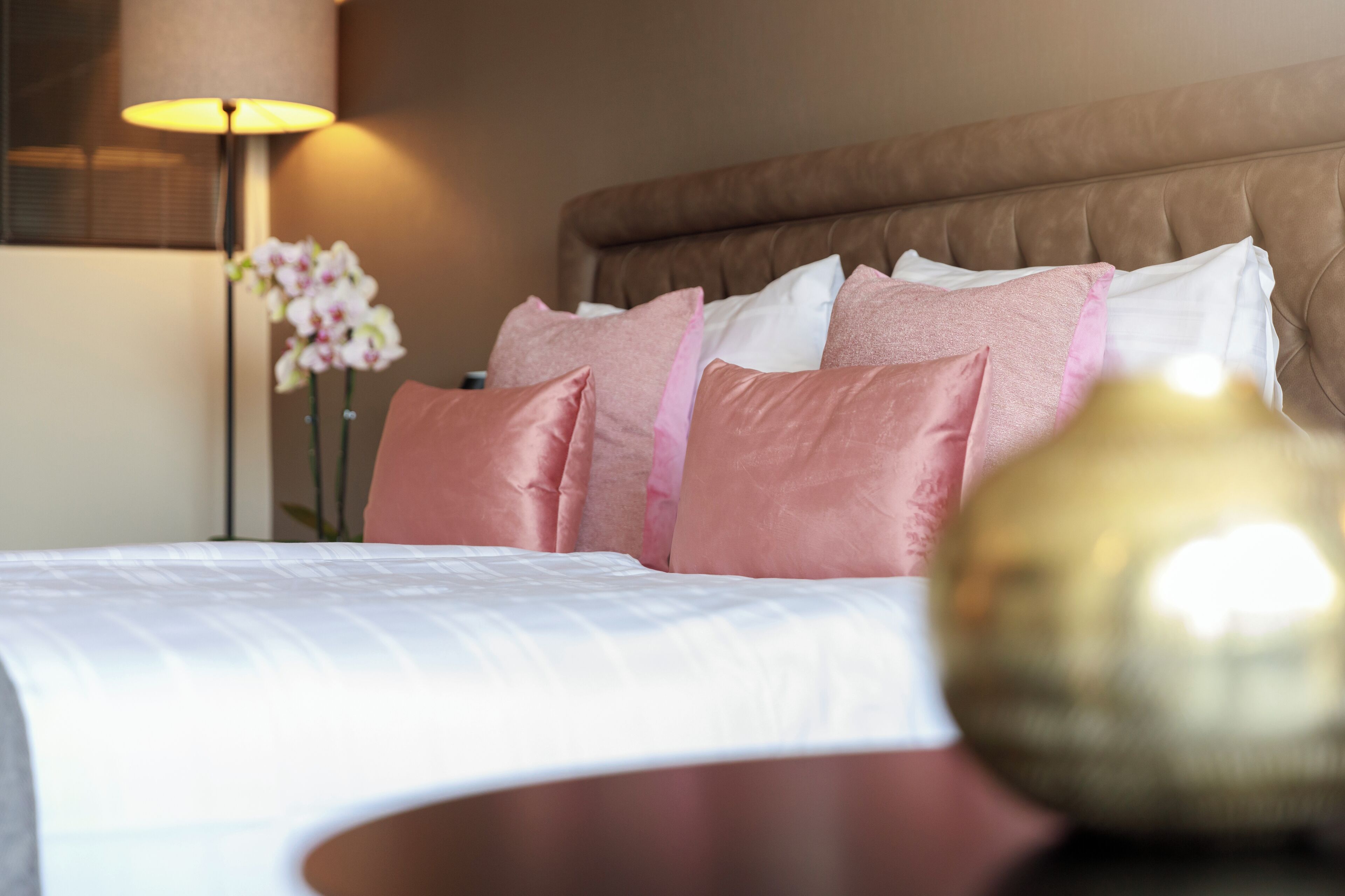 A neatly made bed with pink and white pillows, a bedside lamp, and a vase of flowers.
