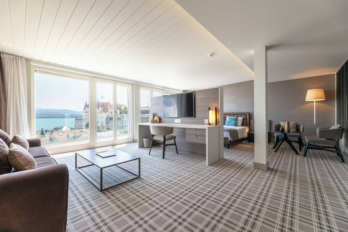 A modern hotel room with a large window offering a city view, a cozy seating area, a flat-screen TV, and a small desk with a chair.