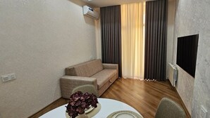 Un espace de vie compact comprenant un canapé, une table à manger avec des chaises, un téléviseur à écran plat fixé au mur et une fenêtre avec des rideaux.