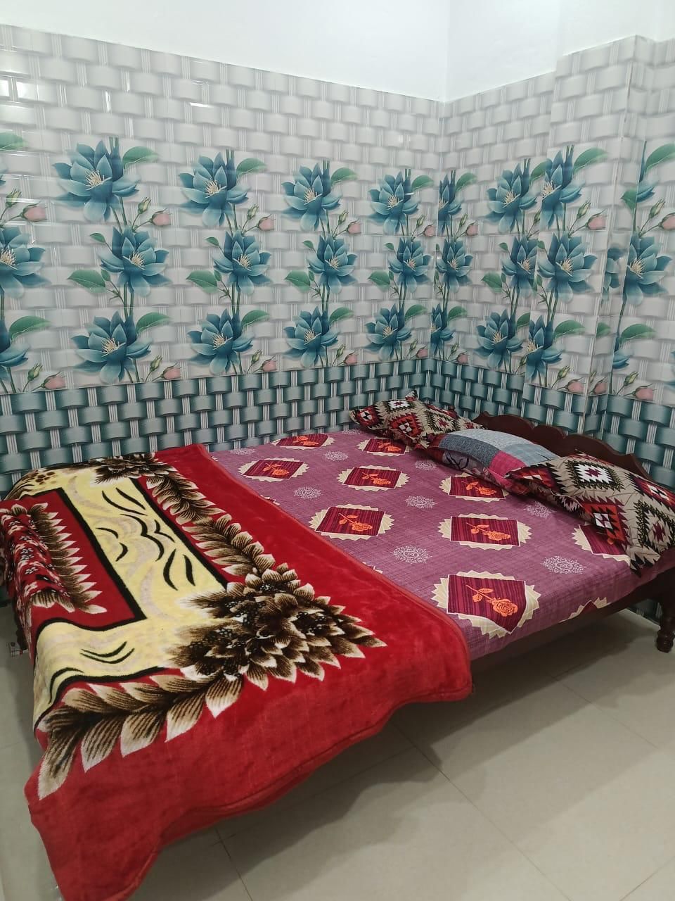 Une chambre avec un lit recouvert d’une couvre-lit rouge et violet à motifs, un coussin décoratif et une couverture à motifs floraux. Un mur de la pièce est recouvert de carreaux décorés de motifs floraux.