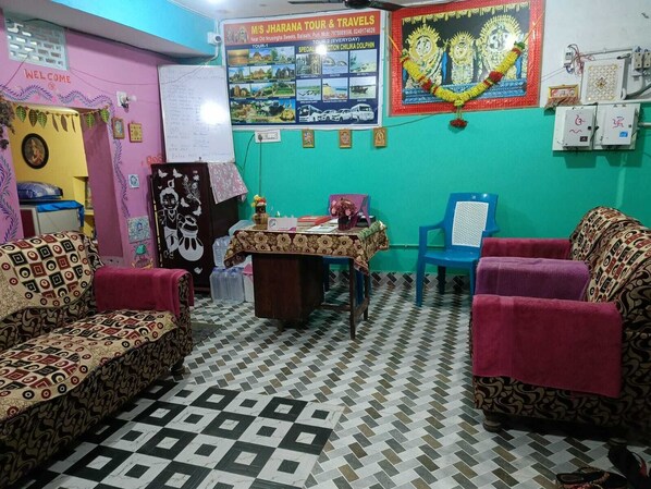 A room with patterned sofas, a small table with a floral tablecloth, a wall-mounted calendar, and a colorful poster. - JHARANA guest house (Puri)